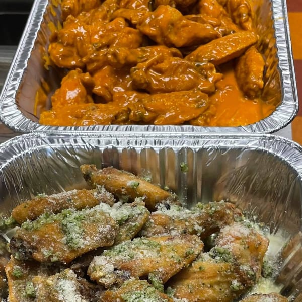 Two foil pans of chicken wings: one with orange buffalo sauce, the other with parmesan and green herbs.